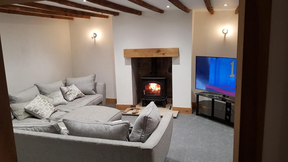 The finished living room — original ceiling beams exposed, the fireplace opened to its stone with an oak mantel and a lit wood burner, and oak underfoot replacing the dark mahogany.